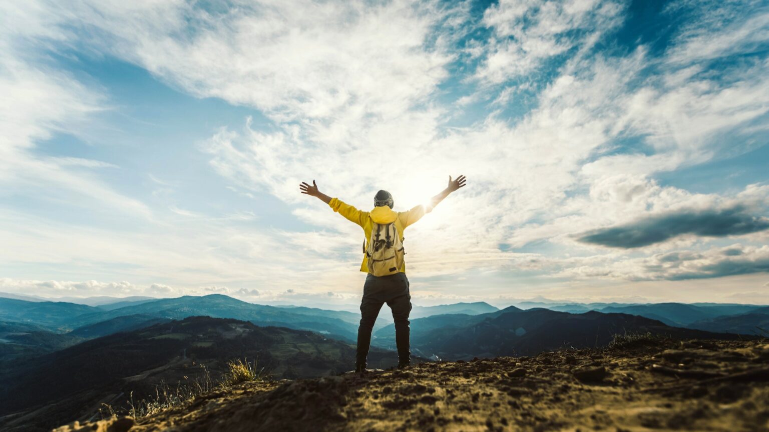 Man traveler on mountain summit enjoying nature view with hands raised over clouds